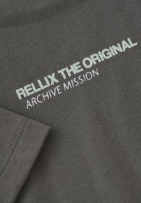 Grauer Stoff mit einer weichen Textur, auf dem in Weiß und hellem Türkis der Text "RELLIX THE ORIGINAL" und "ARCHIVE MISSION" deutlich gedruckt ist.