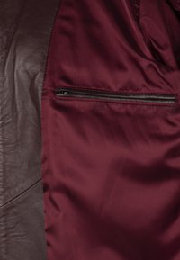 Intérieur d'une veste en cuir brun, avec un revêtement en satin bordeaux profond et une poche zippée. Texture lisse avec des matériaux contrastants.
