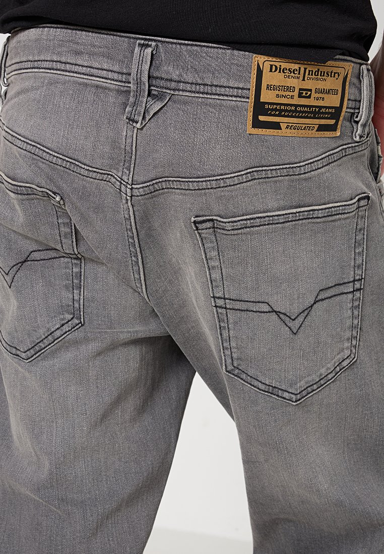 Graue Jeans aus Denim mit zwei Gesäßtaschen, die ein gesticktes V-Muster aufweisen. Ein Lederschild mit Markendetails ist am Bund angebracht.