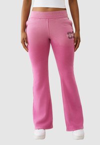 Różowe spodnie dresowe z rozszerzanymi nogawkami, miękkim materiałem i kontrastowymi przeszyciami. Charakterystyczny nadruk na lewej udzie.