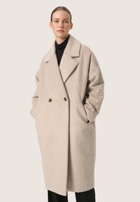Palton bej din lână, cu un design cu două rânduri de nasturi, rever larg și buzunare laterale, având o croială lejeră și o textură moale.