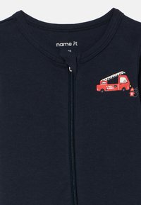 Mornarsko plava pidžama na patentni zatvarač od mekanog pamuka. Ima grafiku crvenog vatrogasnog kamiona na prsima i kontrastni patentni zatvarač. Glatka tekstura po cijelom materijalu.