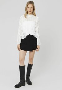 Haut en dentelle blanche à manches longues avec un col en biseau, associé à une jupe mini asymétrique noire ornée de boutons décoratifs. Bottes noires à genoux.