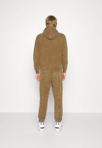 Hoodie in velluto marrone con cappuccio e pantaloni abbinati, caratterizzati da una finitura testurizzata e polsini elastici. Indossato con sneakers bianche.