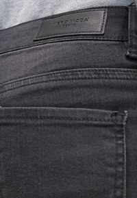 Pantalones de mezclilla negros con una etiqueta de cuero que dice "VERO MODA DENIM" en la pretina, con un diseño clásico de cinco bolsillos y textura sutil.