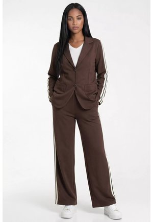 Kobieta stojąca w brązowej marynarce na dwa guziki i dopasowanych szerokich spodniach z białymi pasami bocznymi, białej koszuli i białych sportowych butach.