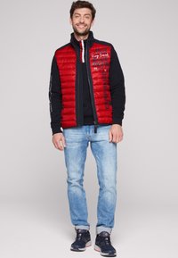 Gilet trapuntato rosso sopra una felpa nera, abbinato a jeans azzurri chiari e sneakers nere con accenti rossi. Testi e loghi sul gilet.