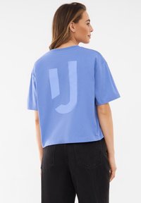 Lys blå cropped t-shirt med korte ærmer og stort lys blåt grafisk print på ryggen, parret med sorte højtaljede bukser.