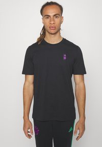Camiseta de algodón negra con mangas cortas, que presenta un pequeño logo morado en el lado izquierdo del pecho y un cuello redondo.