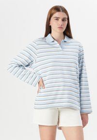 A young woman poses with one hand on her hip, wearing a light blue striped long-sleeve polo and white shorts against a plain background.