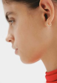 Pendientes de aro dorados con un diseño con picos, que presentan una superficie texturizada. Visibles en un primer plano junto a una vista lateral del rostro de una persona.