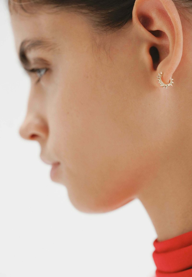 Pendientes de aro dorados con un diseño con picos, que presentan una superficie texturizada. Visibles en un primer plano junto a una vista lateral del rostro de una persona.