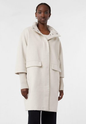 Hellbeige Mantel aus weichem Stoff, mit steilem Kragen, zwei Fronttaschen und gerippten Bündchen. Knopfdetails sind am Hals sichtbar.