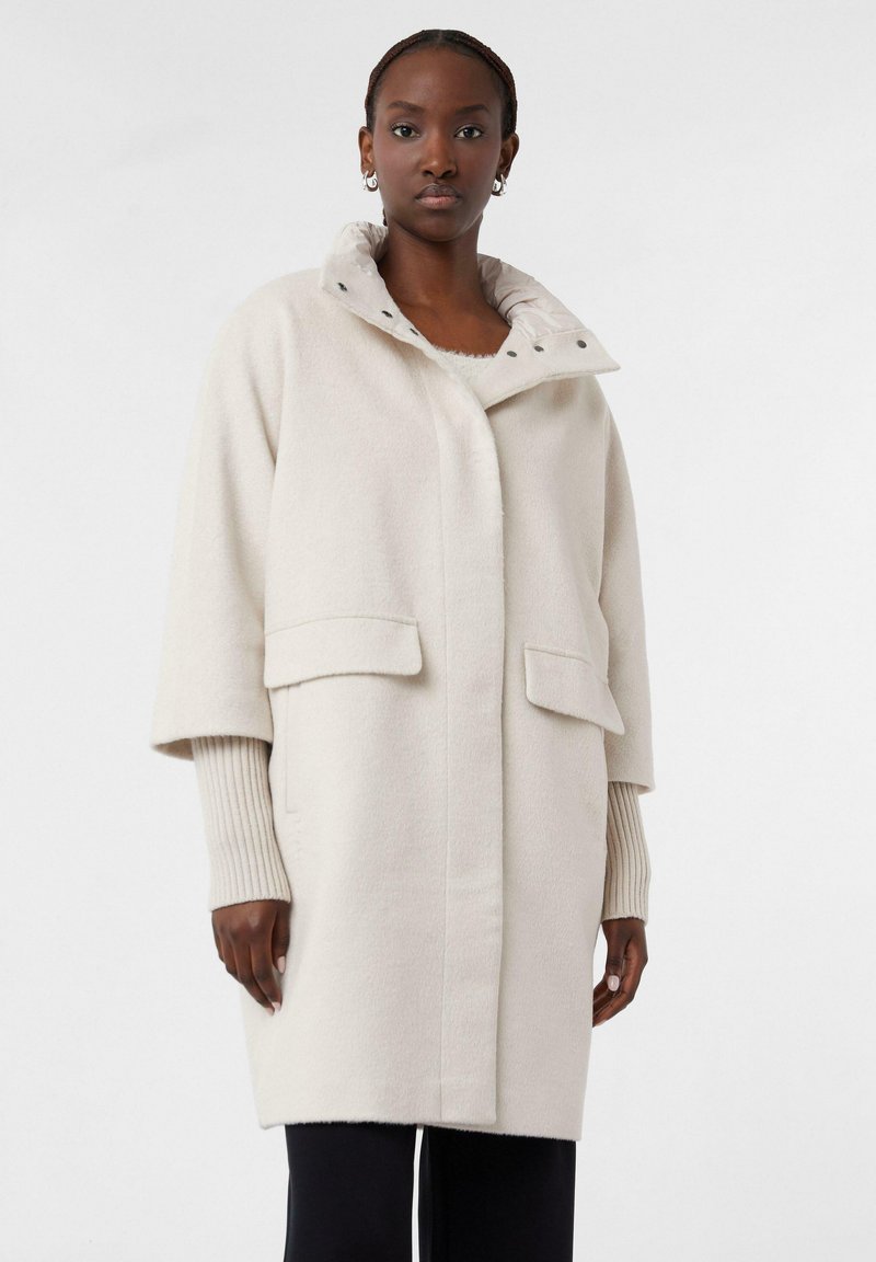 Cappotto beige chiaro realizzato in tessuto morbido, con colletto alto, due tasche frontali e polsini a coste. I dettagli dei bottoni sono visibili sul collo.