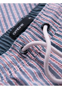 Tissu rayé bleu et blanc avec des accents rouges, cordon blanc passé à travers des œillets noirs portant l'étiquette Ombre Wear sur une ceinture.