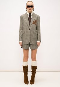 Grau kariertes Sakko mit "M" Stickerei, kombiniert mit passenden Shorts, weißem Hemd, brauner Krawatte und kniehohen, hellbraunen Stiefeln mit strukturierten Socken.