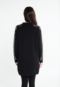 Donna con lunghi capelli scuri che indossa un lungo maglione nero con accenti di paillettes argento sulle maniche e sulle spalle, in piedi su uno sfondo bianco.