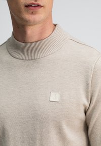 Beige gestrickter Pullover mit einem gerippten Kragen, der ein kleines weißes Logo-Patch auf der Brust trägt. Glatte Textur, lässige Passform.