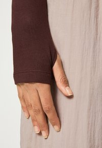 Lång burgunderfärgad ärm med tumhål, åtsittande vid handleden. Armen övergår till ett lätt, texturerat och flödande tyg i en mjuk neutral färg.