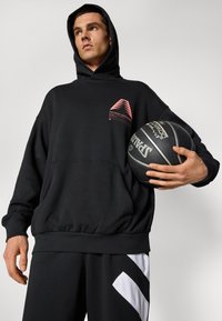 Fekete kapucnis pulóver elülső zsebbel és rózsaszín geometriai logóval. Fekete nadrággal párosítva, amelyen fehér grafikai mintázat található.