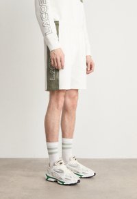 Biezi balti šorti ar zaļu sānu paneļu, uz kura ir "LACOSTE" logo, apvienoti ar baltām un zaļām svītrām zeķēm un daudzkrāsainām sporta apavām.