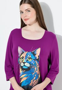 Purppuranvärinen pitkähihaine paita, jossa on värikäs kissagrafiikka. Suunnittelussa on kirkkaita oranssin, sinisen ja vihreän sävyjä yksityiskohtaisilla kasvojen piirteillä.