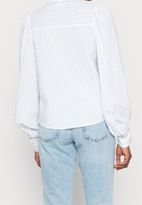 Blouse à rayures blanches et bleu clair avec manches ballon et ourlet arrondi. Caractérisée par un tissu texturé et des rayures verticales au dos.