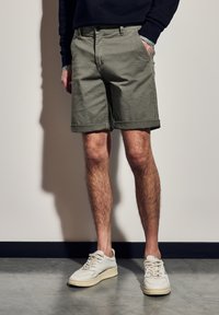 Olivově zelené bavlněné šortky s ohrnutými lemy a bočními kapsami, v kombinaci s bílými teniskami a tmavým svetrem, stojící na texturované podlaze.