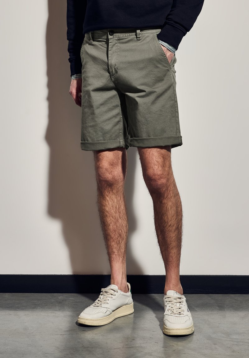 Olivově zelené bavlněné šortky s ohrnutými lemy a bočními kapsami, v kombinaci s bílými teniskami a tmavým svetrem, stojící na texturované podlaze.