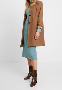 Brun ullkappa med snedskurna slag, svarta knappar; teal ribbat klänning; matchas med bruna krokodilprickade ankelstövlar.