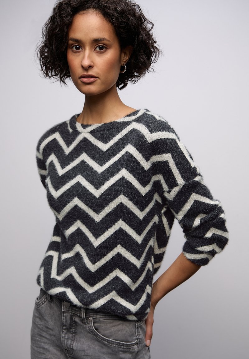 Černobílý svetr s cikcak vzorem, měkkým materiálem, kulatým výstřihem a volným střihem. Modelka má na sobě šedé džíny.