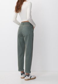 Une femme se tient debout, les mains dans les poches, portant un pantalon taille haute vert olive, un haut à manches longues blanc côtelé, des chaussettes grises et des baskets blanches.