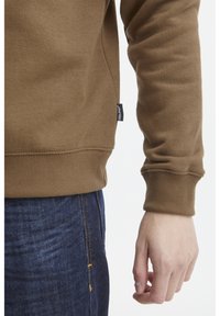 Sudadera marrón con puños acanalados, que cuenta con una etiqueta en la costura lateral. Combinada con jeans de mezclilla azul oscuro, que muestran una textura suave y un diseño sencillo.