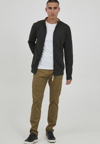 Mørk grå zip-up jakke over en hvit t-skjorte, kombinert med beige bukser. Sorte sneakers fullfører antrekket. Glatte teksturer gjennomgående.