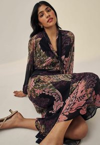 Rochie neagră cu imprimeu floral, cu mâneci lungi, având un model în nuanțe de roz și verde. Țesătura pare ușoară și fluidă, asociată cu sandale nude.