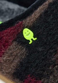 Pantofole infeltrite con una miscela di fibre rosse, marroni e nere presentano un piccolo adesivo di un personaggio verde giocoso sul lato.