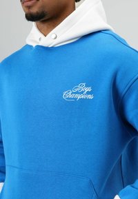Blauwe trui met een witte capuchon, voorzien van een geborduurd "Boys Champions" logo in het wit. Gemaakt van een glad, zacht stof met een comfortabele, losse pasvorm.