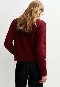 Bordeaux gekropte trui met een gestructureerde afwerking, geribbelde manchetten en zoom, gecombineerd met donkerdenim jeans. Zicht vanuit de achterkant, met lang haar.