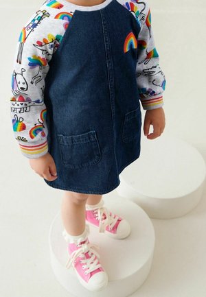 Enfant portant une robe en denim foncé avec des manches à motifs arc-en-ciel et animaux, debout sur des plateformes rondes blanches, baskets roses avec des lacets blancs.