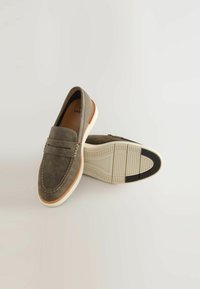 Grå ruskind slip-ons med et mokkasin-design, der har hvide gummisåler og brune detaljer langs kanten. Syninger på overdelen.