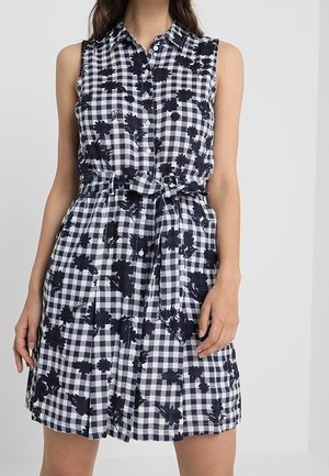 Robe sans manches à carreaux noirs et blancs avec imprimé floral, boutonnée sur le devant, col et ceinture nouée à la taille portée par une femme debout.