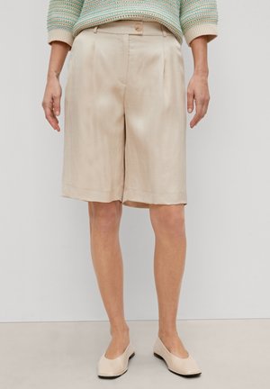 Short beige à jambes larges et longueur genou, avec plis devant, taille haute, fermeture bouton, porté avec des ballerines crème et un haut rayé à manches longues.