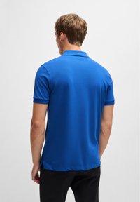 Plava polo majica s kratkim rukavima, izrađena od glatkog materijala. Ima klasični ovratnik i bočne ventile. Stražnji pogled prikazuje prilagođeni kroj.