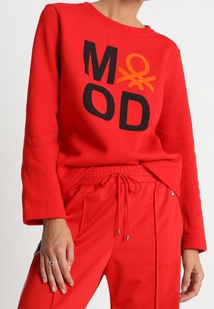 Rotes Langarmoberteil mit "MOD" und orangefarbenem Logo, kombiniert mit roten Kordelzughosen, getragen von einer stehenden Person mit Ringen an den Fingern.