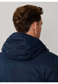 Chaqueta con capucha azul marino de textura suave; cuenta con capucha fruncida y detalles de herrajes en contraste. Vista desde atrás.