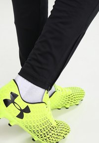 Jasně žluté fotbalové boty s texturovaným zigzag vzorem, kombinované s černými sportovními kalhotami s zipy na kotnících a bílými ponožkami.