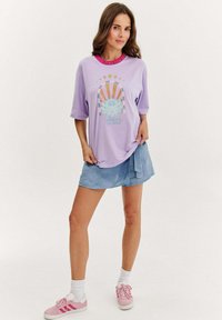Fialové oversized tričko s designem lotosu, obsahující vrstvenou grafiku v pastelových barvách. Spárováno s světle modrou sukní a růžovými teniskami.