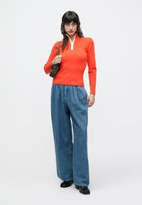 Haut rouge à manches longues avec col zippé, associé à un pantalon en denim bleu ample. Le modèle tient un petit sac vert. Chaussures noires. Fond blanc.