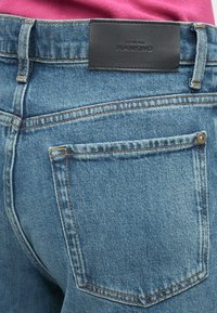 Blugi din denim cu o spălare albastru deschis, având cusături portocalii, un buzunar dreptunghiular în spate și o etichetă de brand din piele neagră pe talie.