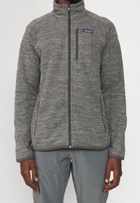Veste en polaire grise avec un col haut, fermeture éclair à l'avant, poches latérales et tissu texturé. Présente un petit logo sur la poitrine gauche.